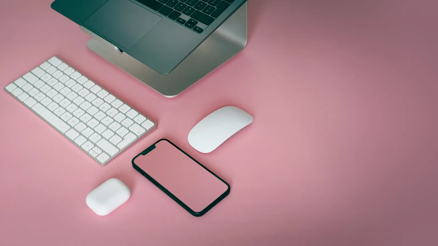 Pembe zemin üzerinde dizüstü bilgisayar, klavye, fare ve telefondan oluşan sade masa